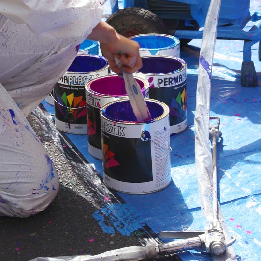 Close up image of an artist wearing white overalls kneeling on a blue tarp with one hand holding a wooden paint mixer mixing one colourful pot of paint amongst many